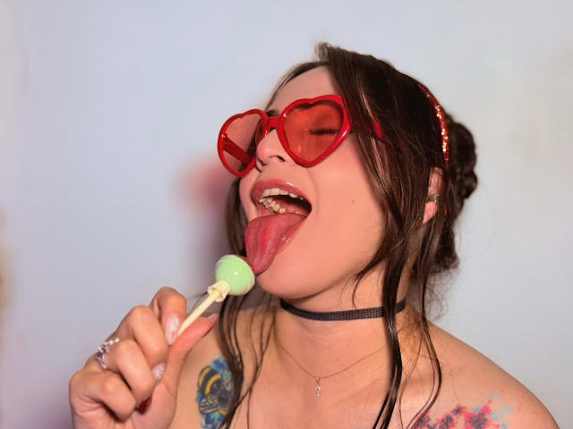 ElinaJhonson with red heart-shaped glasses, tongue out, licking a green lollipop, black choker, tattoo on right shoulder, messy brown hair, light skin, and a nose ring.