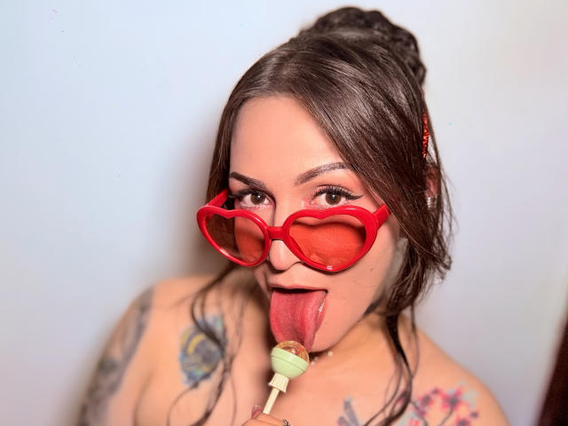 ElinaJhonson with heart-shaped red glasses, tongue out, licking a green lollipop, tattoos on her shoulders, dark brown hair in a bun, looking at the camera.