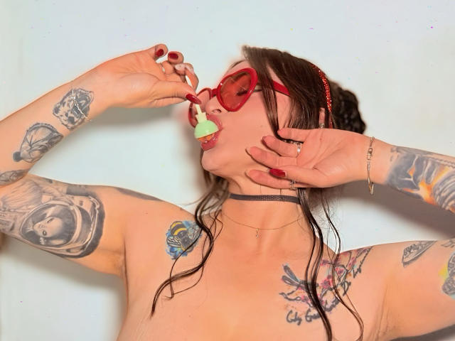 ElinaJhonson with red heart glasses, holding a green lollipop to her mouth, has multiple tattoos, wet brown hair, and is topless, wearing a black choker and red nails.