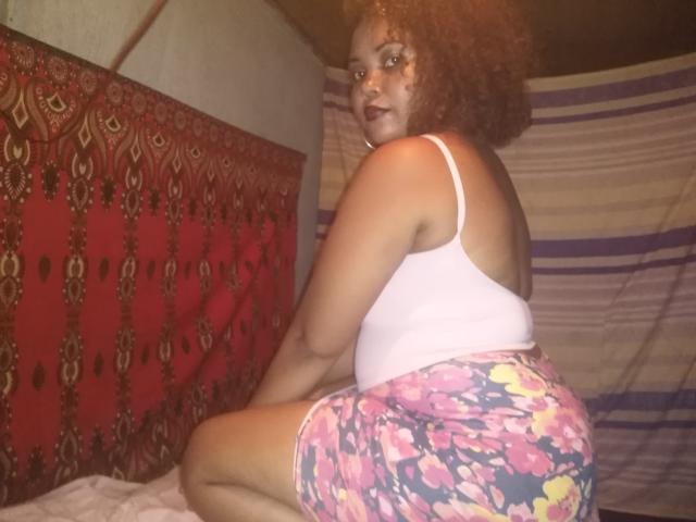 DjaoSimila69 has curly brown hair, medium brown skin, wears a pink tank top, and floral shorts, kneeling on a bed with a red patterned pillow. Her ass is visible.