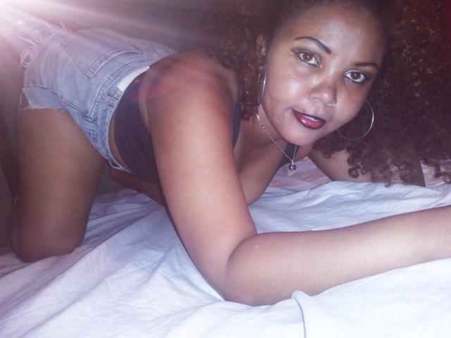 DjaoSimila69, with curly brown hair and hoop earrings, on all fours, wearing denim shorts and a black top, showing her ass and looking at the camera with a smile.