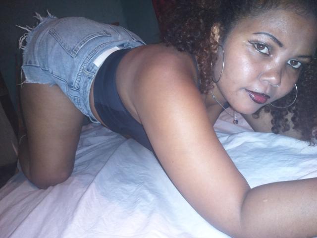 DjaoSimila69 on all fours, wearing denim shorts and a black tank top, looking at the camera with curly brown hair, hoop earrings, and a slight smile. Her ass is up.