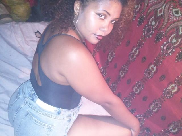 DjaoSimila69 with curly brown hair, dark skin, wearing a black tank top and light blue denim shorts, sitting on a bed, looking over her shoulder, showing her ass.