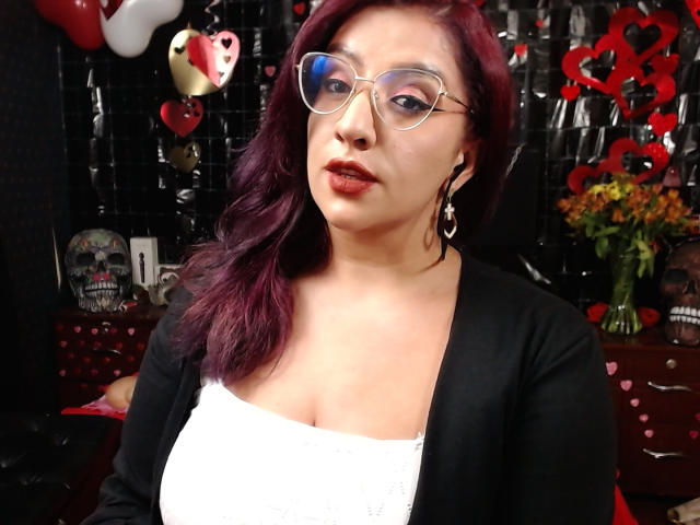 AndreaSharick with purple hair, glasses, red lipstick, white top, black cardigan, showing her tits, black background, skull decor, heart balloons, flowers, and a dark, shiny wall.