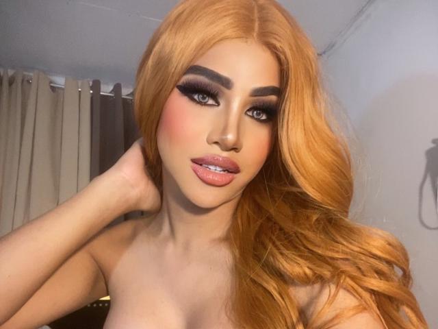 MilkyAnora, con cabello largo y naranja, maquillaje intenso, labios rosados y cejas marcadas, tocándose la cabeza con una mano, mostrando sus hombros desnudos y sus pequeños senos..