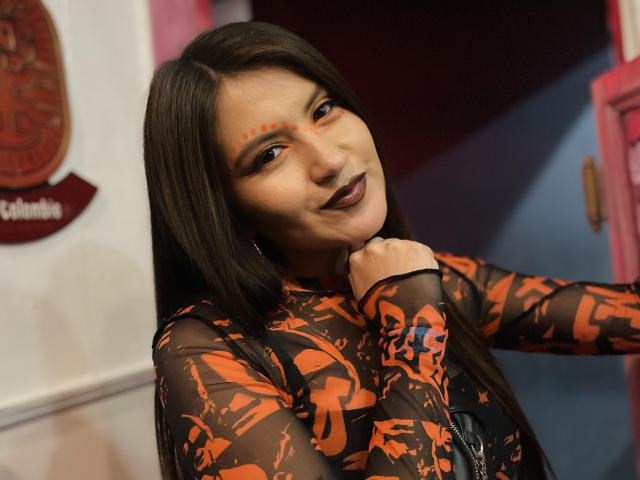 AmelieMuller with dark lipstick, orange face paint, and long brown hair in a sheer black and orange patterned top, holding her chin, smiling. Boobs, tits, pussy, and ass are not visible.
