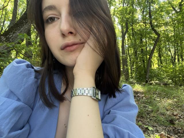 LilieFlower a un visage mignon avec des cheveux bruns qui couvrent un œil, elle porte une chemise bleue à manches bouffantes et une montre argentée à la main, et repose sa joue..