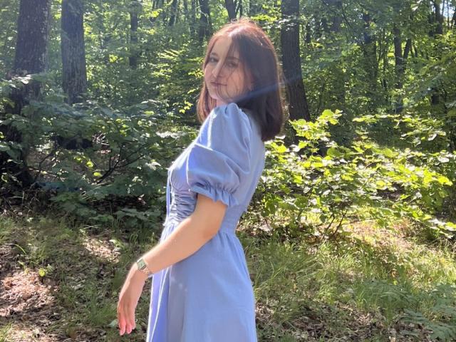 LilieFlower, avec une poitrine de taille moyenne, portant une robe bleu clair, debout dans une forêt baignée de soleil, regardant en arrière avec un léger sourire, cheveux bruns, arrière-plan de feuilles vertes..