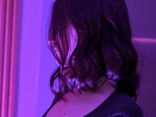 LilieFlower, avec ses cheveux noirs et ondulés qui lui cachent les yeux, porte un haut noir, éclairée par des lumières violettes et roses. Son visage est partiellement ombragé, avec un regard subtil et mystérieux..
