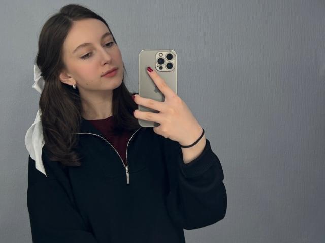 LilieFlower with brown hair, wearing a black zip-up hoodie, red shirt, and white ribbon, holding a silver iPhone, taking a mirror selfie with red nails.