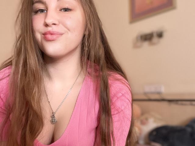 Dooris with long brown hair, pink ribbed top, showing cleavage, smiling, wearing a silver necklace with a butterfly pendant, in a casual indoor setting.
