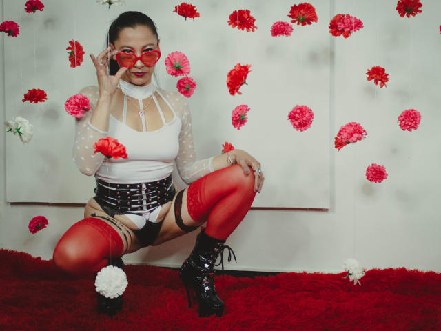 Noemi, in una posa elegante, esegue uno squat indossando collant rossi, una maglietta bianca a pois, un corsetto nero e tacchi alti, tenendo in mano occhiali rossi, con fiori rosa e bianchi e un tappeto rosso..