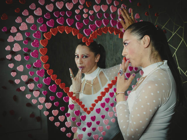 NoemiPrettiest with pouty lips, wearing a sheer white polka dot dress, kissing her reflection in a mirror framed by red and pink heart-shaped decorations. She's got a ring on.