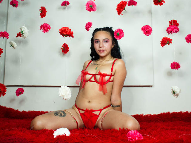 KateBrunet, with red lingerie and a flower in her hair, sits on a red shaggy carpet, surrounded by floating flowers, showing her tits, pussy, and tattoo on her thigh.