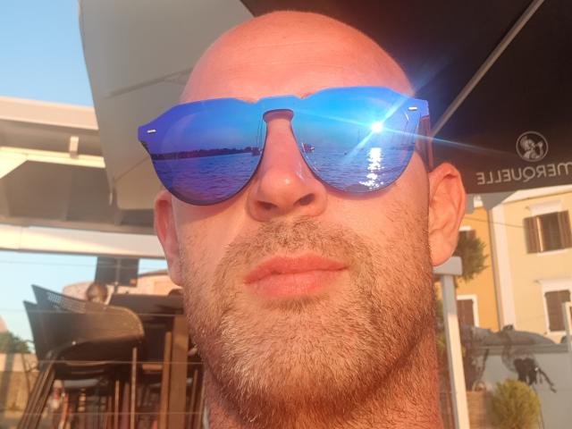 Rox69s has a bald head, a beard, and is wearing blue reflective sunglasses with a sun flare in the upper right. Rox69s has a serious expression.