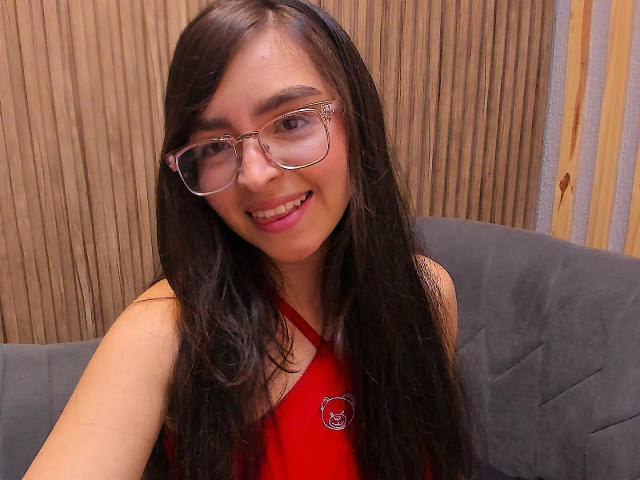 Eva McAllister, com cabelos pretos longos, óculos e uma blusa vermelha de alça, sorrindo para a câmera, mostrando seu rosto fofo e seios pequenos, sentada em um sofá cinza..