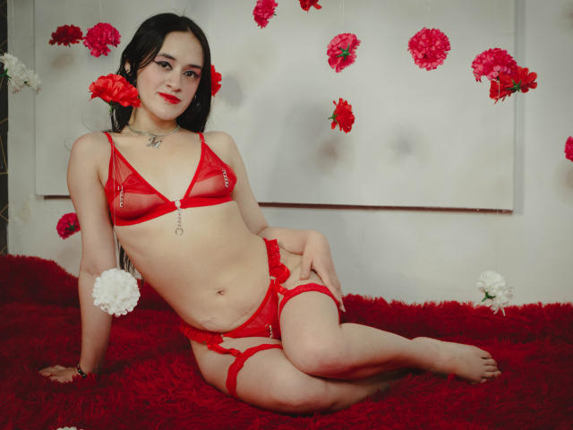 NellyCris with red lingerie, red flowers in hair, sitting on red shaggy carpet, wearing a red bra with straps and matching panties, showing her tits and ass.