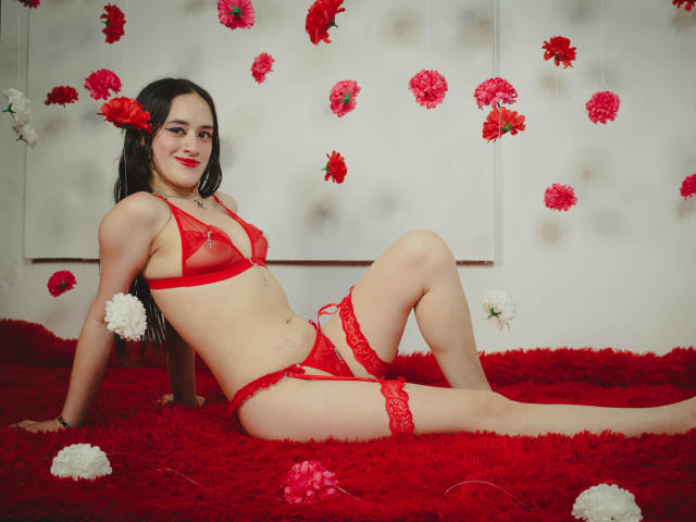 NellyCris with red lingerie, red flowers in hair, lying on red shag carpet, red garter on thigh, smiling, red lipstick, red bra, red panties, small tits, pale skin.