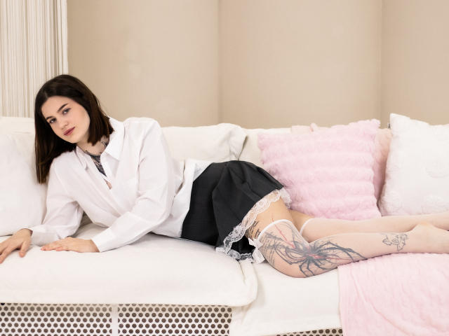 NicoleKissYou, with black bob and white blouse, lies on white couch, wearing black skirt, white stockings, and showing large tattoo on thigh, legs up, pink pillow behind.