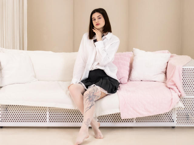 Nicole, met donker haar, een wit shirt, een zwarte rok en witte sokken, zittend op een witte bank, met een getatoeëerd been zichtbaar, met een roze deken en witte kussens..