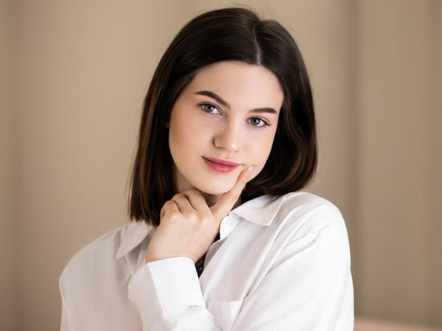 NicoleKissYou, met kort bruin haar, draagt een witte blouse, heeft haar vinger op haar kin, glimlacht zachtjes, draagt roze lippenstift, en haar borsten zijn niet zichtbaar..