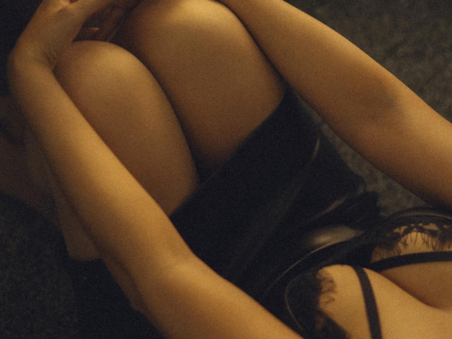 KarinaCecillia's knees are together, her arms wrapped around them. She's wearing a black lace top, her bare legs and arms are visible. The image has a warm, dark tone.