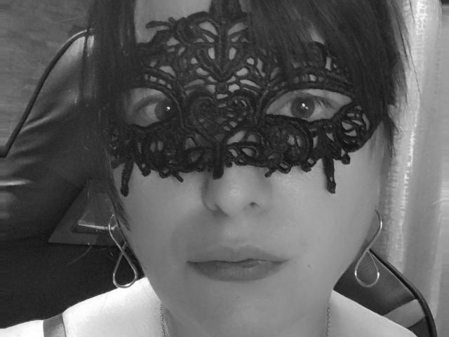 Black and white photo of LeylaaXxx with lace mask covering eyes, dark hair, hoop earrings, and neutral expression, showing nose and lips. She's wearing a thin necklace.