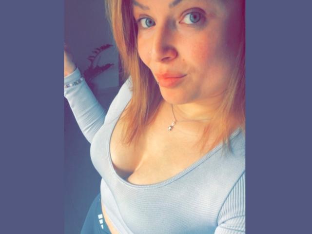 CherieCherry mit blauen Augen, heller Haut und blonden Haaren, trägt ein hellgraues, geripptes Crop-Top, das ihre Brüste zeigt, steht mit einem leichten Lächeln und einer Hand an der Wand..