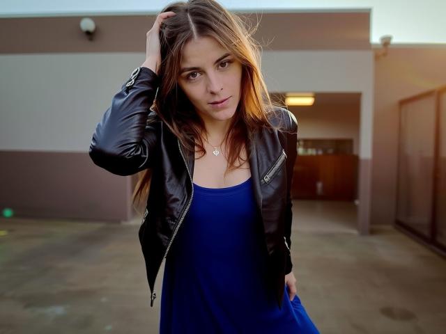 GlamMegi mit braunen Haaren, trägt eine schwarze Lederjacke und ein blaues Kleid, Hand im Haar, blickt in die Kamera, kleine Brüste, mittelgroßer Hintern, weiches Licht, im Freien, leichtes Lächeln..