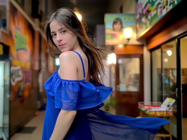 GlamMegi with long brown hair, blue off-shoulder dress, standing in a narrow alley, looking back with a slight smile, subtle makeup, light skin, soft light, night setting.