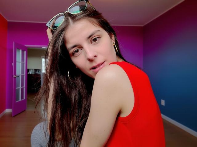 GlamMegi with long brown hair, red sleeveless top, green-framed glasses on head, gold hoop earrings, looking at camera, in room with pink and blue walls.