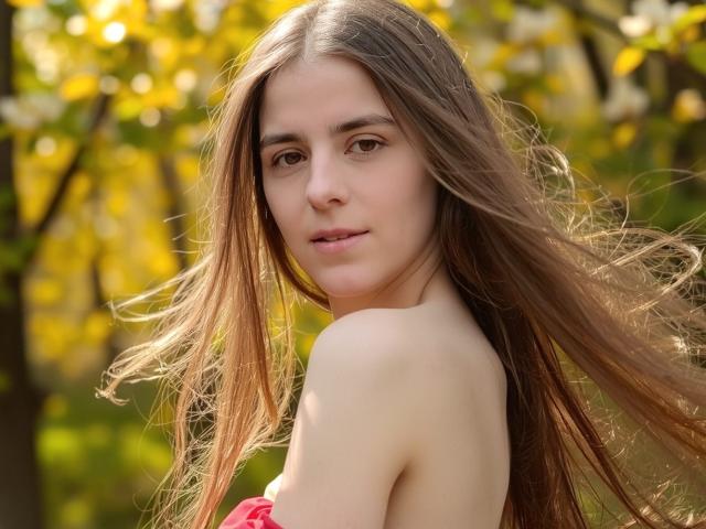 GlamMegi met lang bruin haar, onbedekte schouders, een roze top, een zachte glimlach en een lichte huid, staat in een zonnig bos. Haar bruine ogen kijken zachtjes naar de camera..