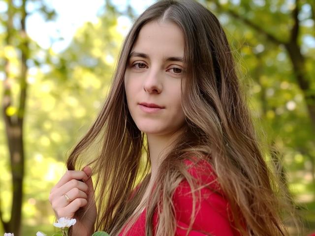 GlamMegi mit langen, braunen Haaren, die ein rotes Hemd trägt, eine weiße Blume hält und in die Kamera schaut, mit einem sanften Lächeln, hellgrünen Augen, im Freien, sonnig, entspannt, süß..