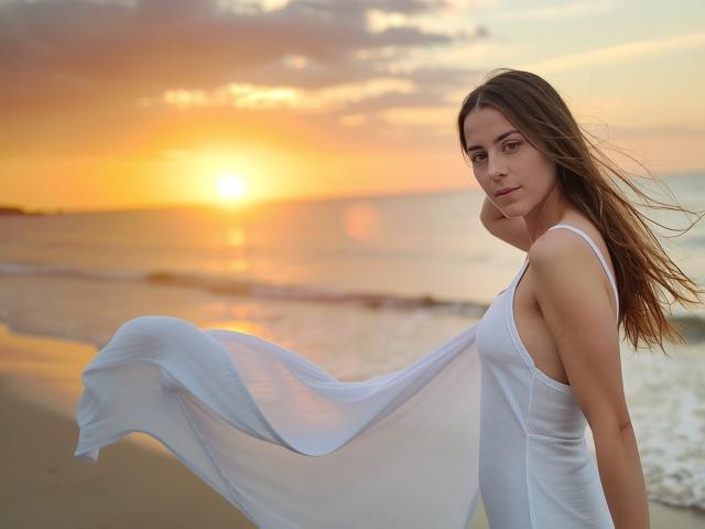 GlamMegi, con i suoi lunghi capelli castani, indossa un abito bianco e fluente, e si trova su una spiaggia al tramonto. Il suo abito ondeggia nel vento, e i suoi seni sono visibili in modo discreto, conferendole un aspetto sensuale..