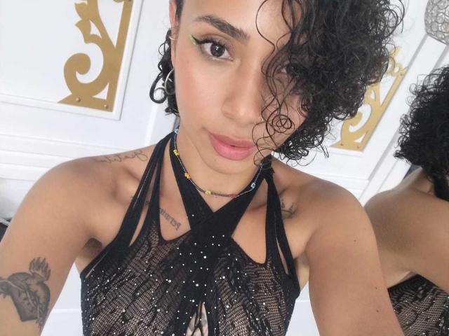 SilvanaXOwen has curly black hair, green eyeliner, and wears a black, sheer, sparkly top with cross straps, showing her small tits and tattoos, looking at the camera.