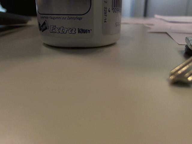White cylinder with "Extra Nuvor" label, sitting on a grey, slightly reflective surface. Key with a metallic, shiny finish is partially visible on the right.