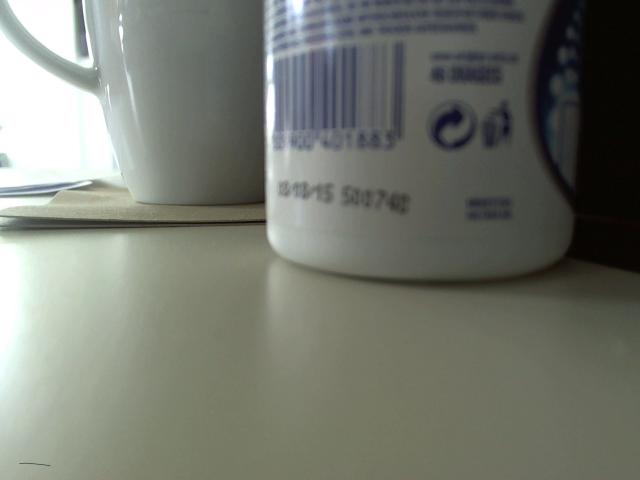 White ceramic mug with blue barcode on a light-colored surface, slightly blurry, next to a similar white ceramic mug on the left. Olegator-hot's ass is a mug.
