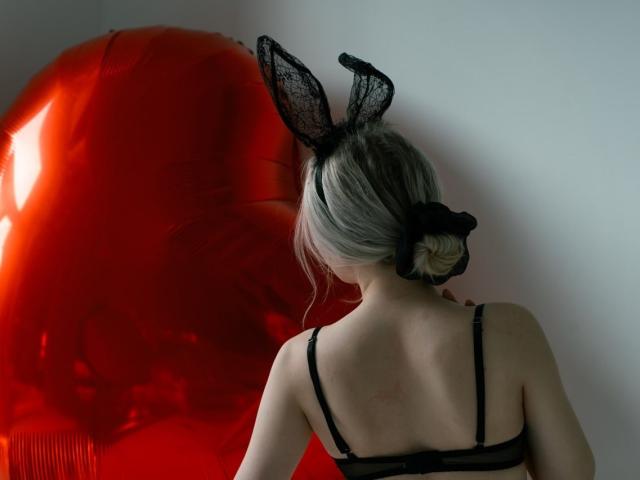 BeatricaKlein with blonde hair in a bun, wearing black lace bunny ears and black bra, standing in front of a large red balloon. Her back is to the camera.