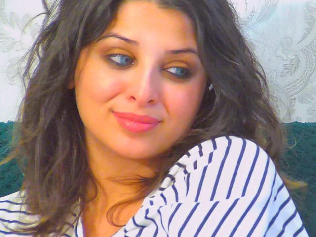 ScarletOra with wavy brown hair, blue eyes, and pink lips, wearing a striped white and blue shirt, smiling slightly, looking to the side. Boobs not visible.
