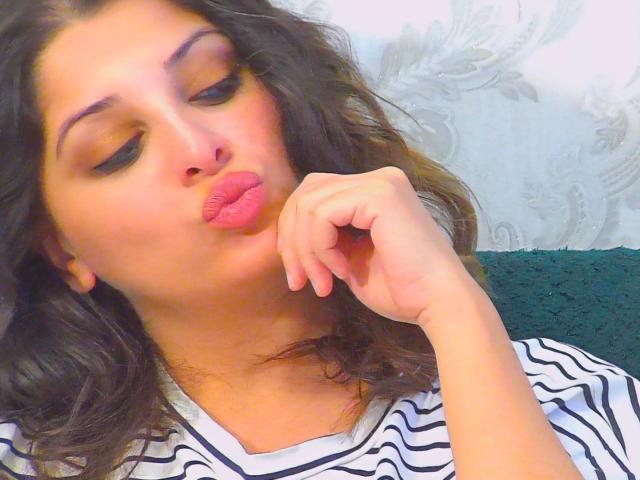 ScarletOra with pouty pink lips, brown wavy hair, and striped shirt, touching her chin with her right hand, looking down with slightly closed eyes.