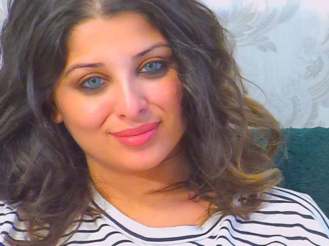 ScarletOra has curly brown hair, blue eyes, pink lipstick, and a slight smile, wearing a white and black striped shirt. Her tits are not visible.