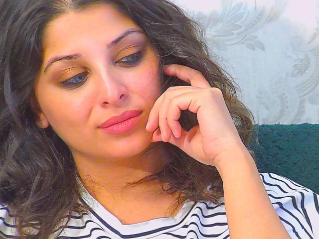 ScarletOra with wavy dark hair, light brown eyes, pink lipstick, and striped shirt, resting her chin on her hand, looking slightly down with a subtle smile.
