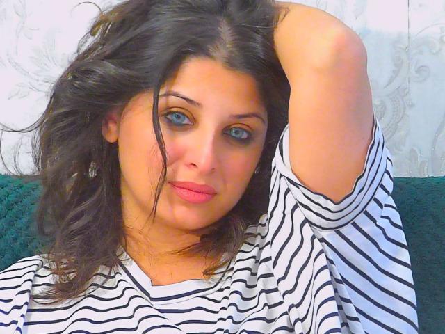ScarletOra has blue eyes, pink lipstick, and is wearing a striped shirt, with one hand in her messy black hair. Her tits are not visible.