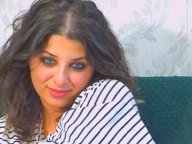 ScarletOra has wavy dark brown hair, blue eyes, and a slight smile. She's wearing a striped shirt, with her tits partially visible. Her pink lips are slightly parted.