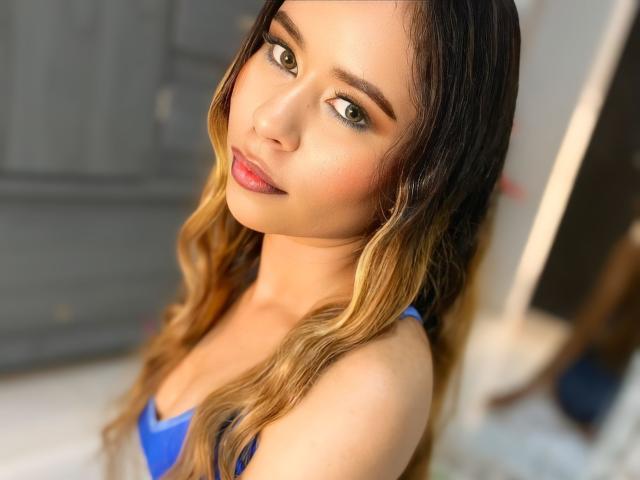 RoxyRosee has blonde ombre hair, green eyes, and a cute face with pink lips. She's wearing a blue top, showing her boobs, and looking seductively at the camera.