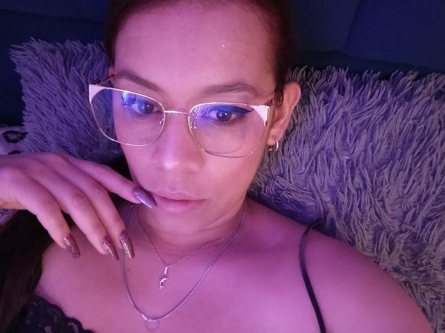 Melodyn with glasses, purple nails, and a necklace, lying down, showing her face and shoulder, looking at the camera, wearing a black strap, purple lighting.
