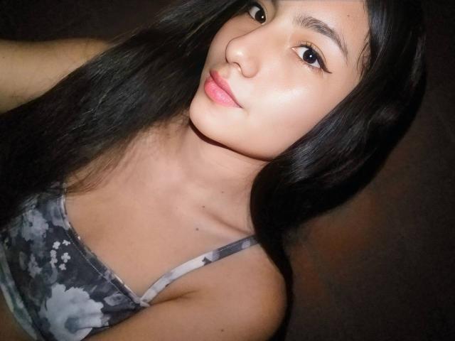 AnieDemond has long black hair, brown eyes, and a cute face with pink lips. She's wearing a gray floral bra, showing her small tits. Her skin is light brown.