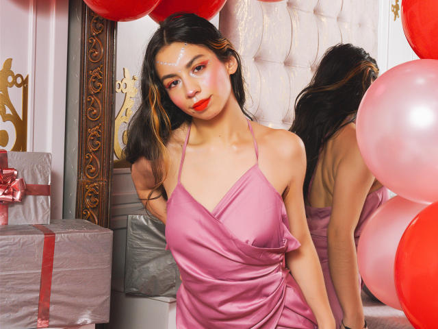 KatherinJohns with shiny pink dress, red lipstick, and glowing face, standing by a mirror with red and pink balloons, showing her bare shoulders and cute ass in the reflection.