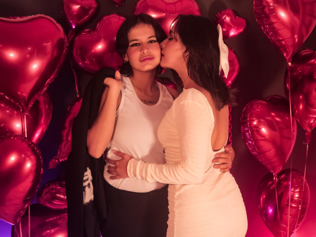 AnneAndSophia with dark hair, white top, and black pants, standing, holding black jacket, being kissed on the cheek by AnneAndSophia in white dress, surrounded by pink heart balloons.