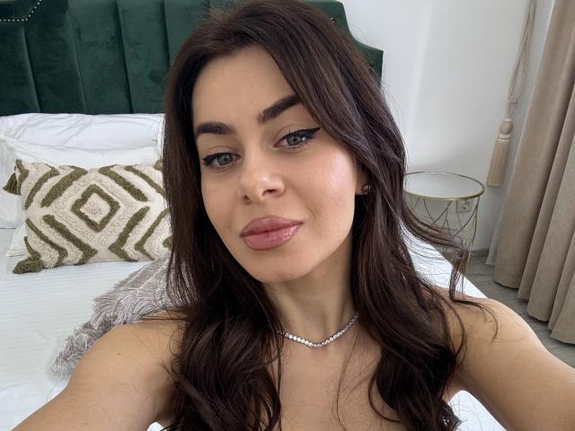 LauraineX with dark brown hair, green eyes, and pink lips, wearing a pearl necklace, showing her upper body and smiling at the camera. Boobs, green headboard in background.
